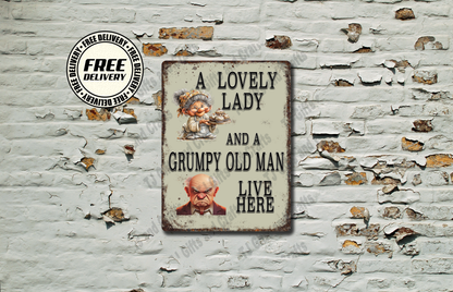A rectangular vintage-style metal sign with a distressed cream background and black text reading 'A LOVELY LADY AND A GRUMPY OLD MAN LIVE HERE,' displayed against a white brick wall with peeling paint. The sign includes small illustrated portraits and a circular 'FREE DELIVERY' badge appears in the upper left.