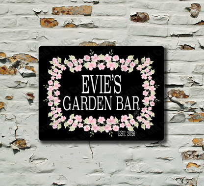 A rectangular black sign with a pink floral border and the text 'EVIE'S GARDEN BAR' mounted on a distressed white brick wall.