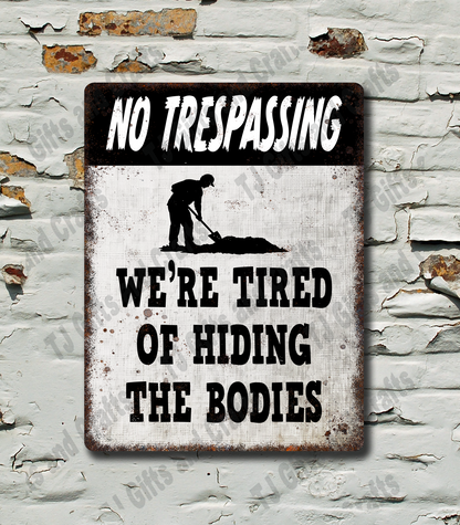 A rectangular metal novelty sign with a distressed white background and black text reading 'NO TRESPASSING' at the top and 'WE'RE TIRED OF HIDING THE BODIES' at the bottom, with a black silhouette of a person digging. The sign is mounted on a white brick wall with peeling paint.