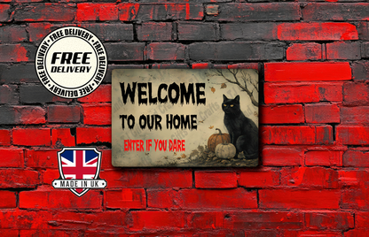 A rectangular decorative sign with a distressed beige background, featuring the text 'WELCOME TO OUR HOME ENTER IF YOU DARE' in black and red, alongside an illustration of a black cat, pumpkins, and autumn leaves. The sign is displayed against a red and black brick wall backdrop, with promotional badges for 'FREE DELIVERY' and 'MADE IN UK' on the left.